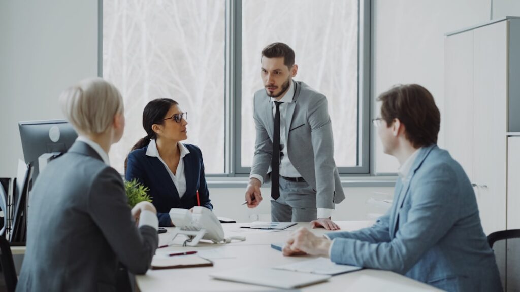 Cuatro personas en reunión laboral. Hombre de pie explicando, traje gris. Otros sentados escuchando.
